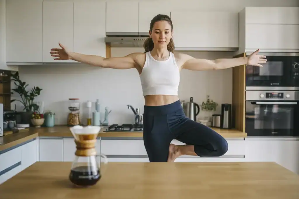 A women standing on one leg practicing balancing body in a modern kitchen while waiting for her cofee to brew. promoting moderate movements at home ahowcasing how to lose weight without exercise.